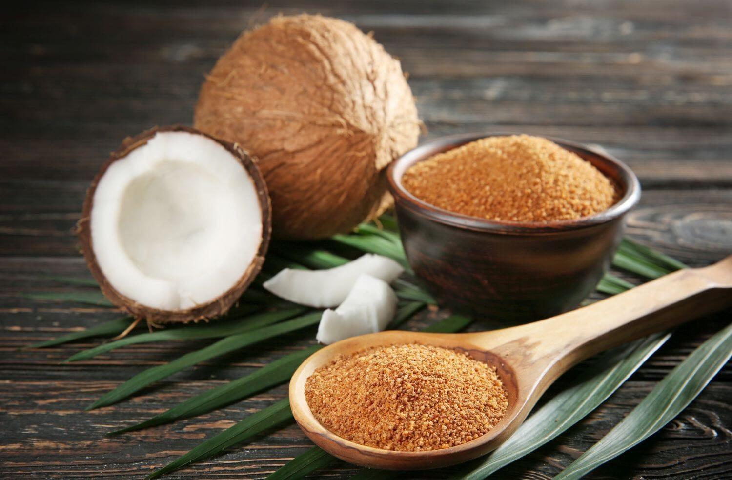 This is a picture of a coconut and a half a coconut with palm sugar in a brown bowl and a wooden spoon on a wooden background.