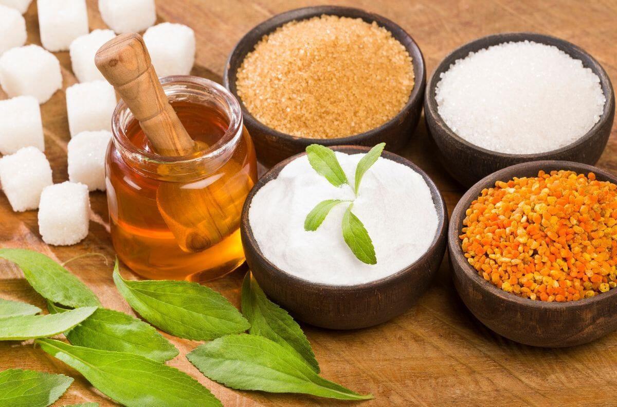 This is a picture of different sweeteners, including honey in a glass jar, coconut sugar in a brown bowl and sugar cubes all on a wooden tabletop.