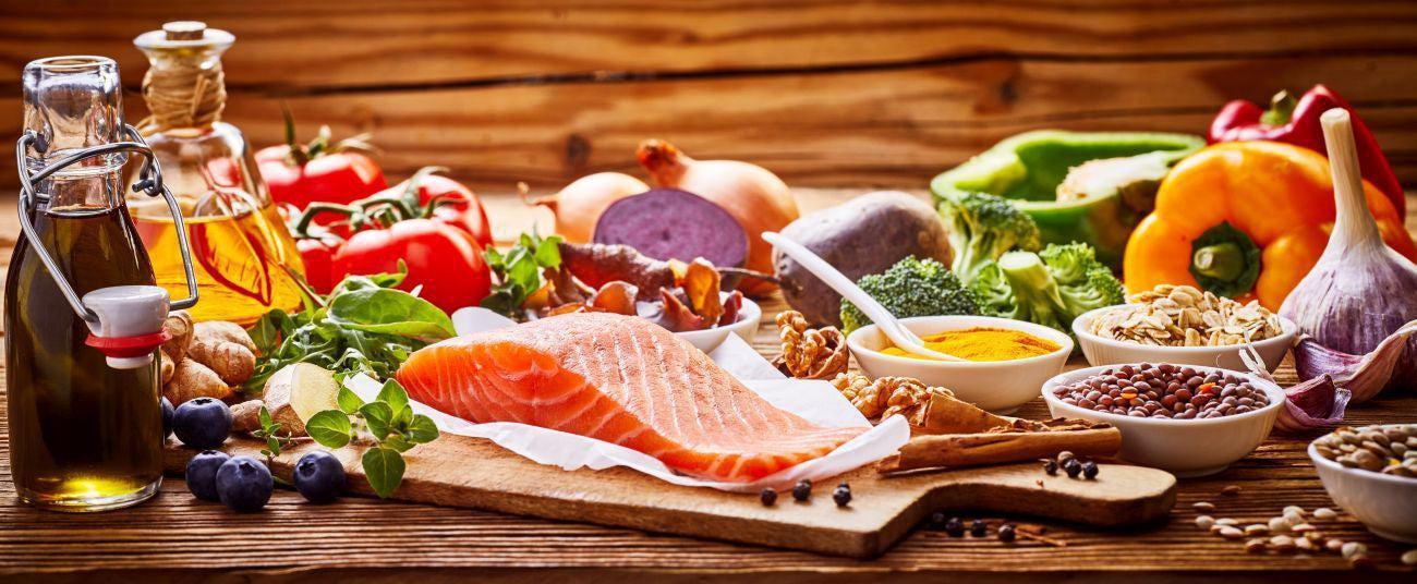 This is a picture of many Mediterranean diet foods such as a salmon filet, olive oils, blueberries, tomatoes on the vine, bell peppers, broccoli, onions, and turmeric powder on a wooden table in glas bottles,
