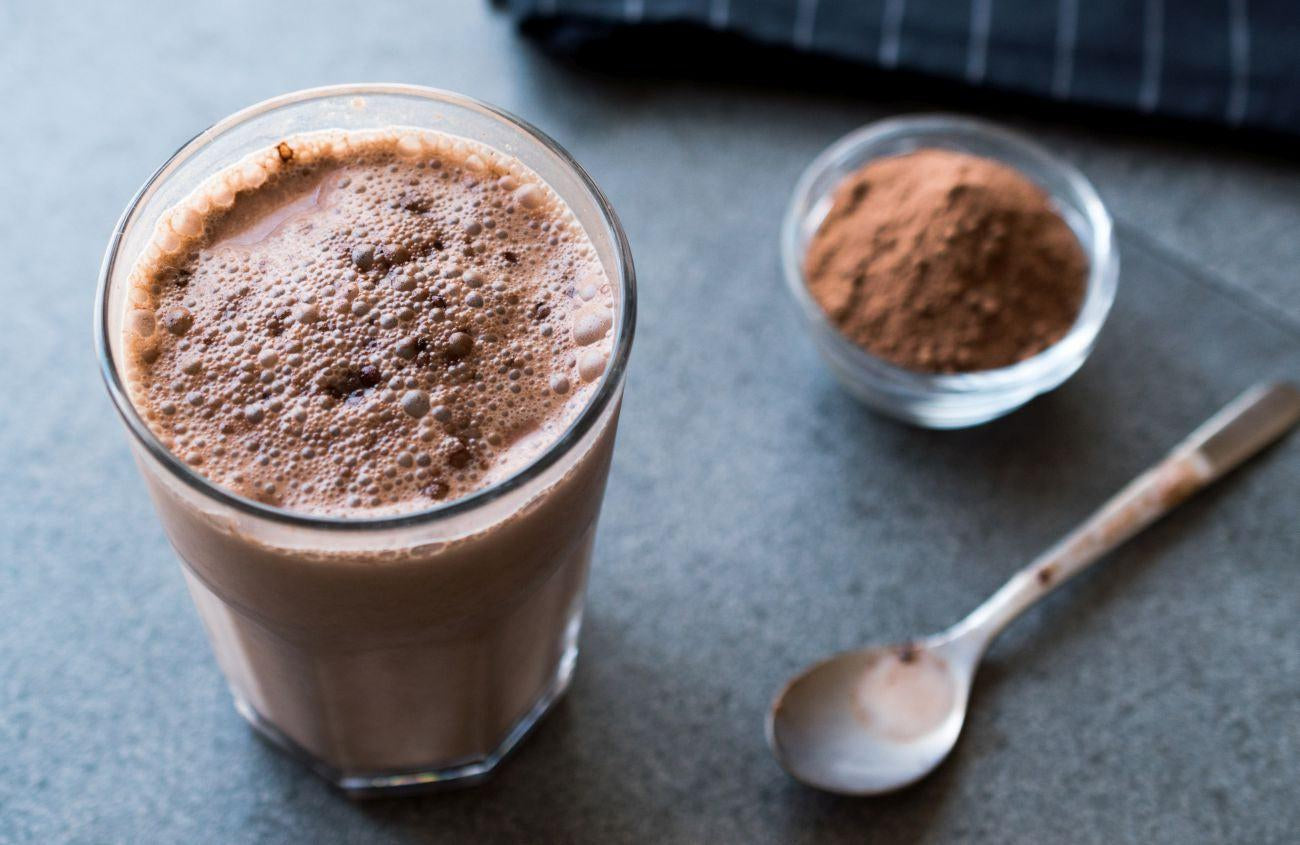 This is a picture of a glass of chocolate milk and powder with a spoon on a dark table top
