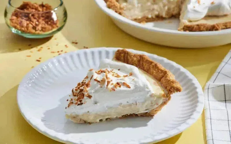 This is picture of a slice of our Coconut Cream Pie with a fork in the middle on a white plate with a white background.