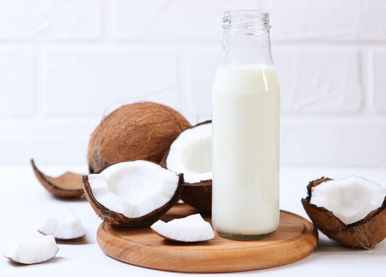 This is a picture of coconut milk in a glass bottle with coconut peices around it on a wooden board on a white background.