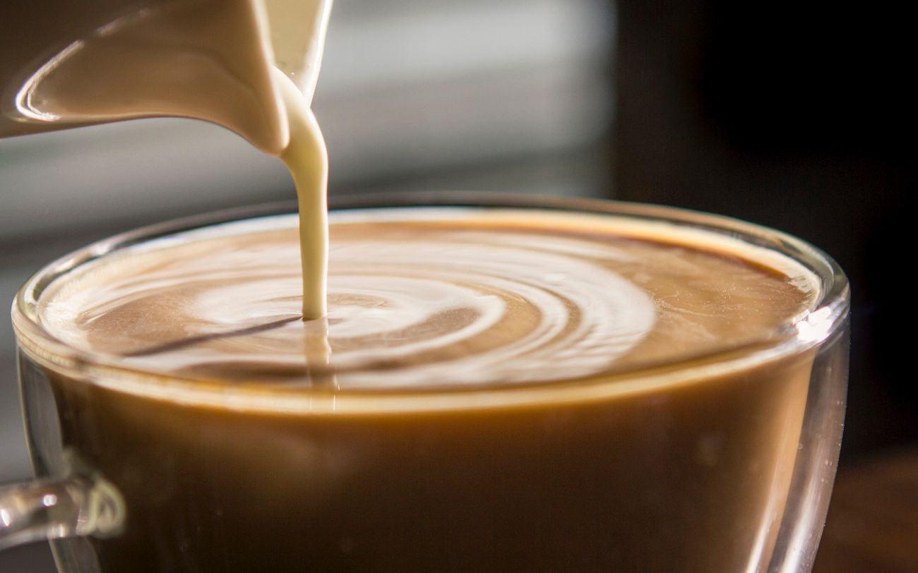 This is a picture of a cup of coffee in a glass mug, with cream being poured into it, with a blurred background