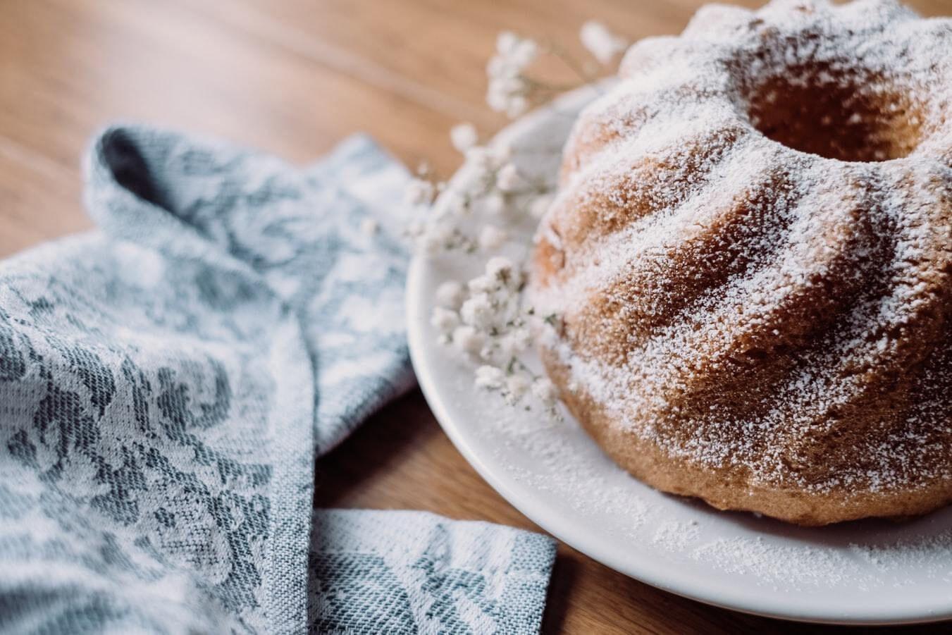 This is a picture of our Coconut Pound Cake with powdered sugar on top sitting on a white plate with a blue napkin on a wooden table top.