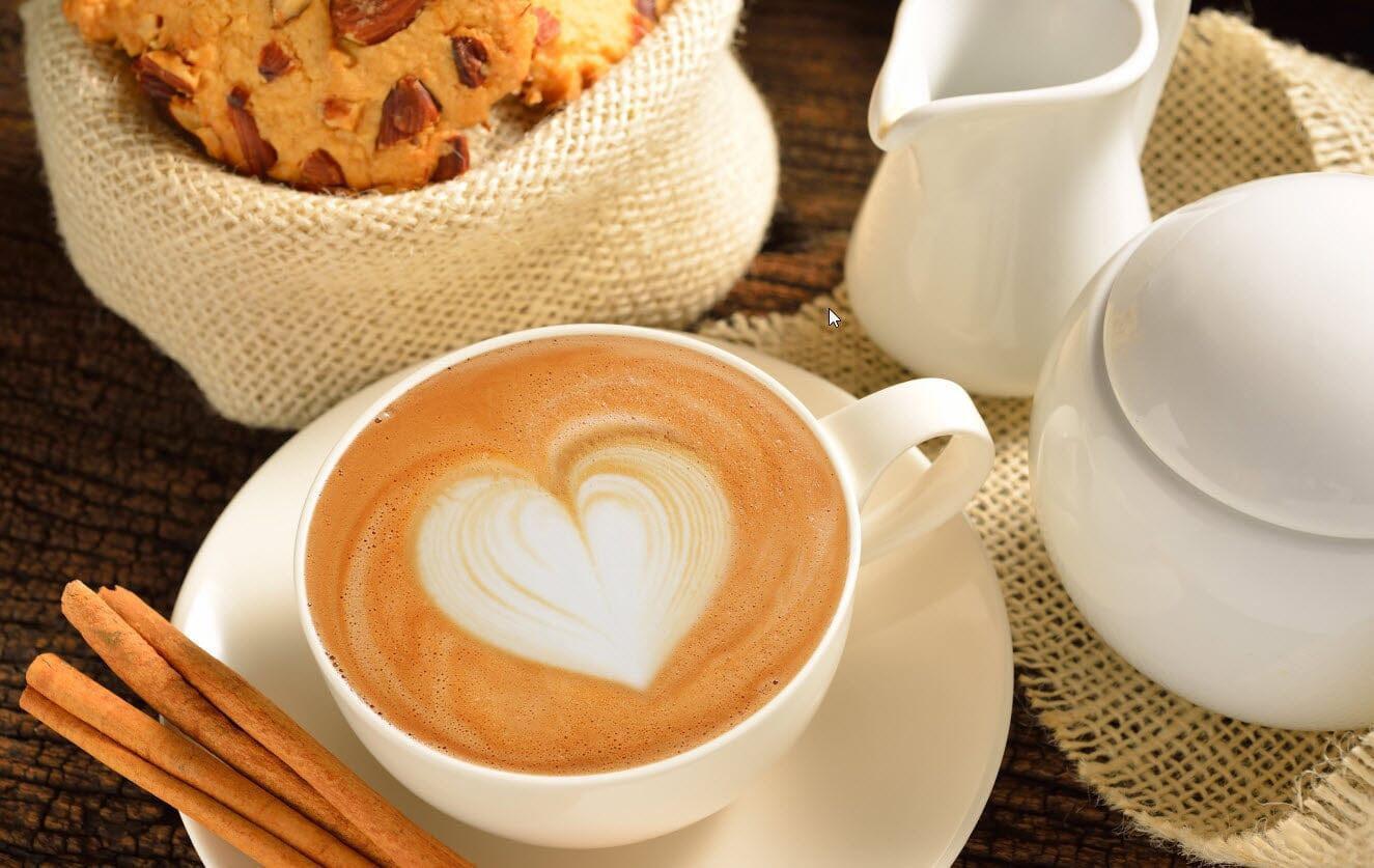 This is a picture of a coffee drink in a white cup with a heart design in the coffee, with cinnamon sticks in the cup saucer
