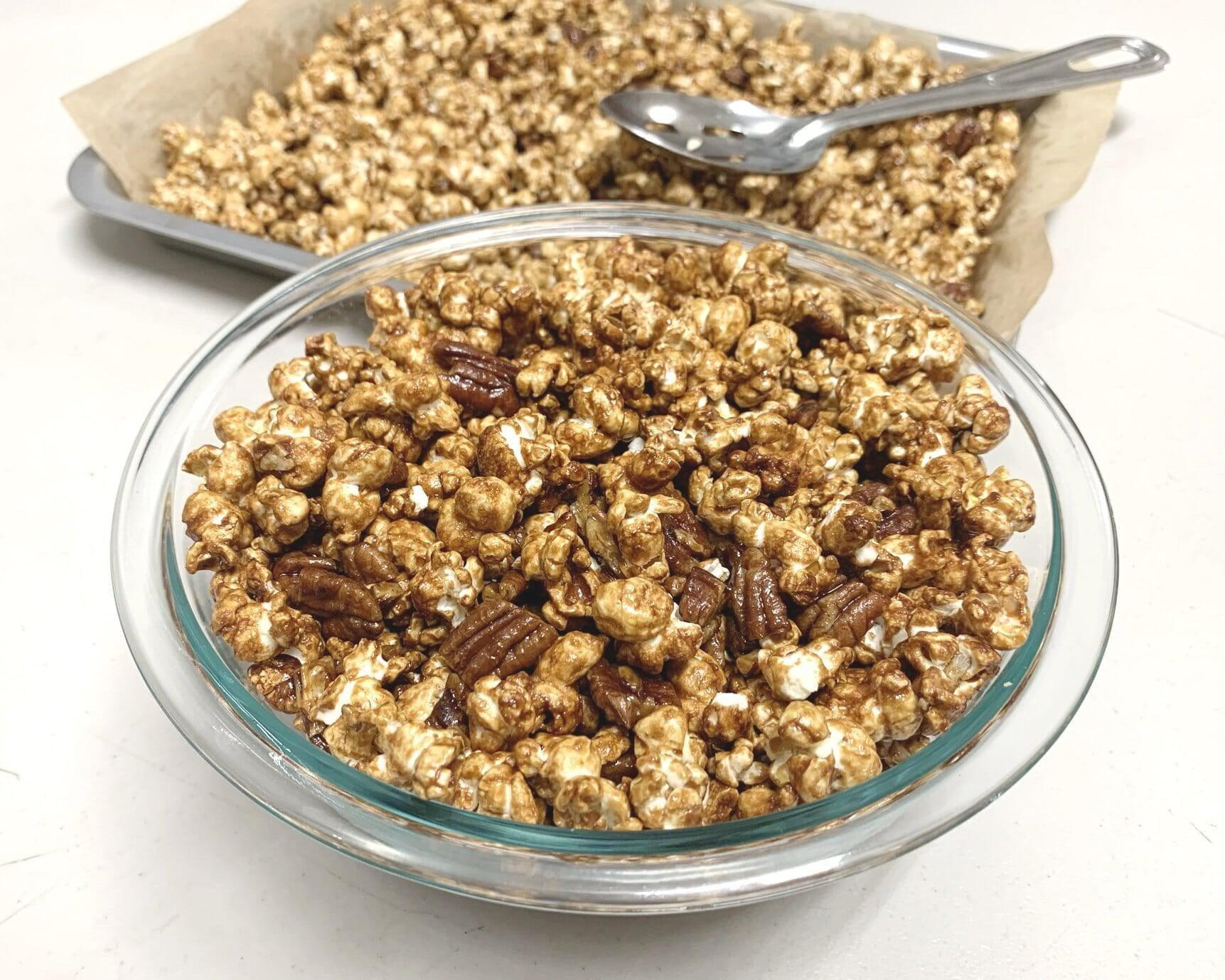 This is a picture of caramelized popcorn in a glass bowl with a pan of popcorn behind it with a silver serving spoon, on a white background