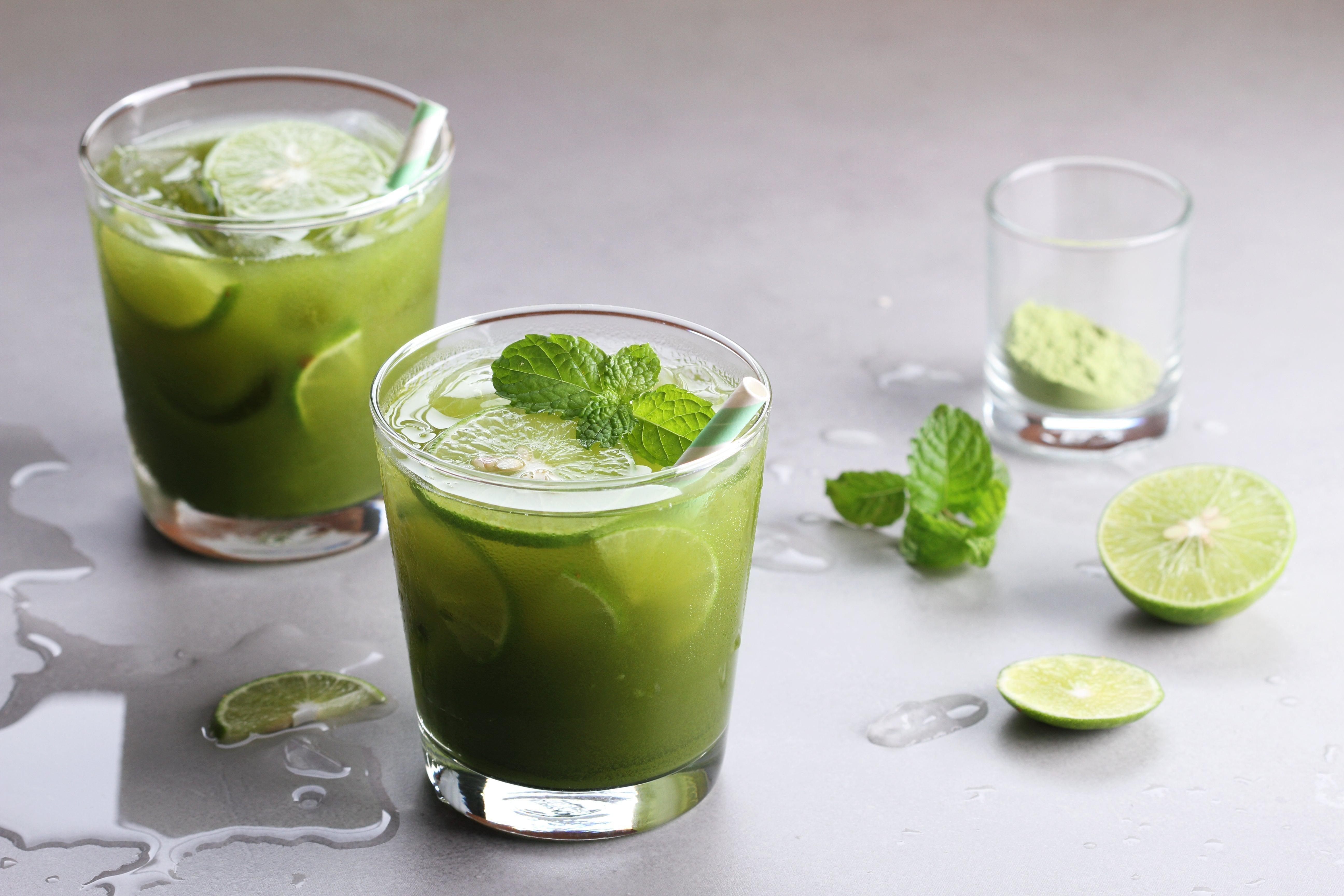 This is a picture of two glasses of Matcha Green Tea Lemonade with slices of lime on a grey table top.