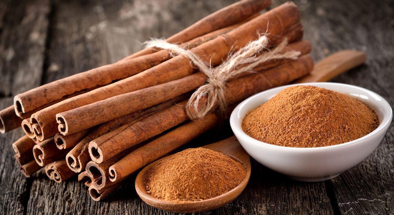 This is a picture of a bundle of fresh cinnamon sticks next to a wooden spoon filled with cinnamon powder and a white bowl with cinnamon powder on a wooden table