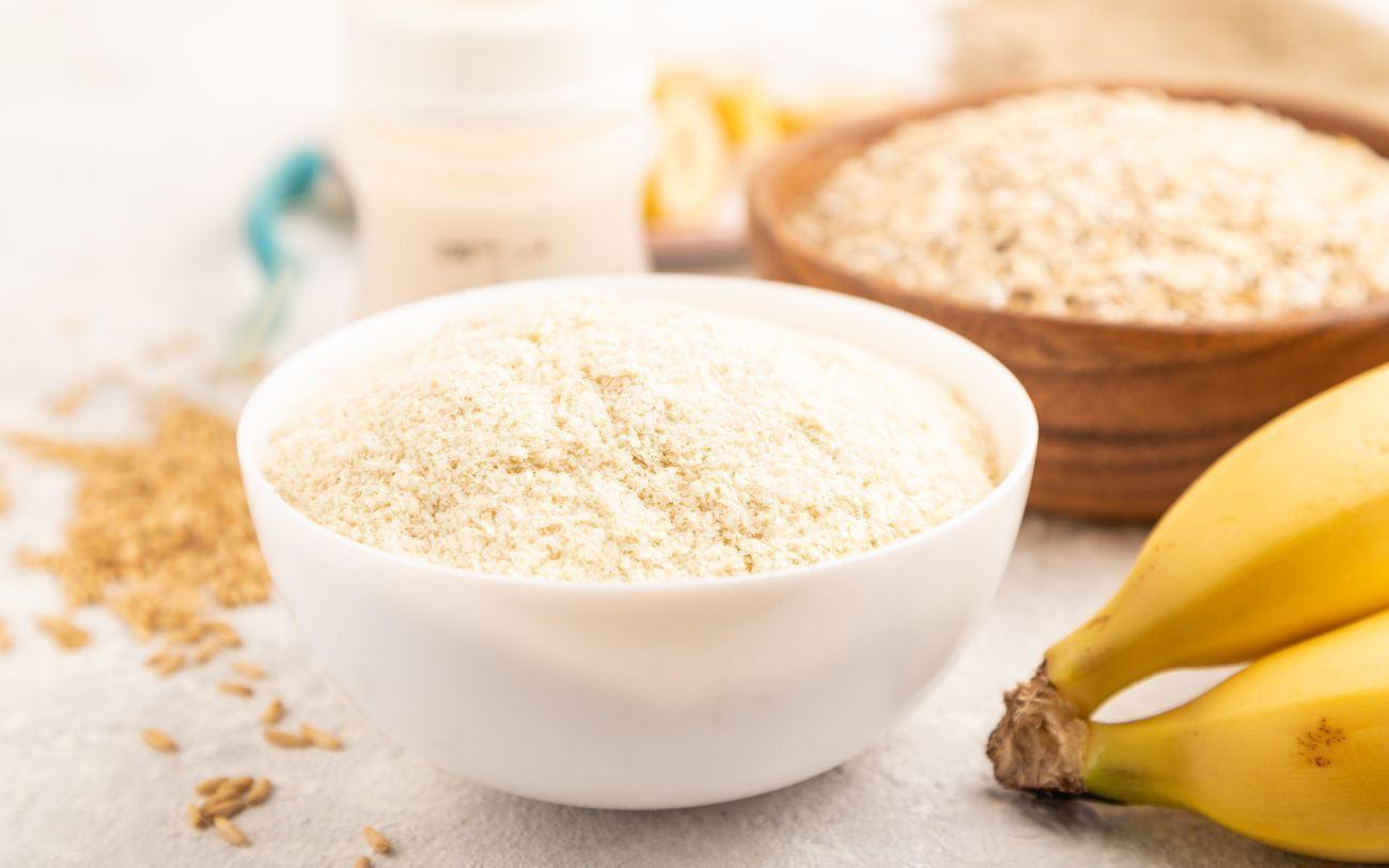 This is a picture of powdered milk and a jug of milk, bananas and a wooden bowl of oatmeal on a white table top.