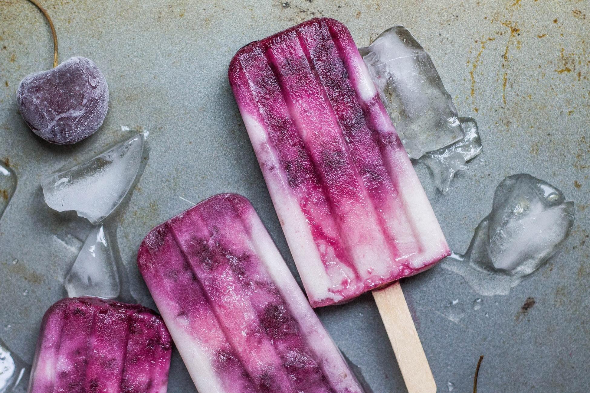 This is a picture of 3 red and white colored superfood popsicles with ice cubes laying on a greyish colored stone backround.Superfood Popsicles