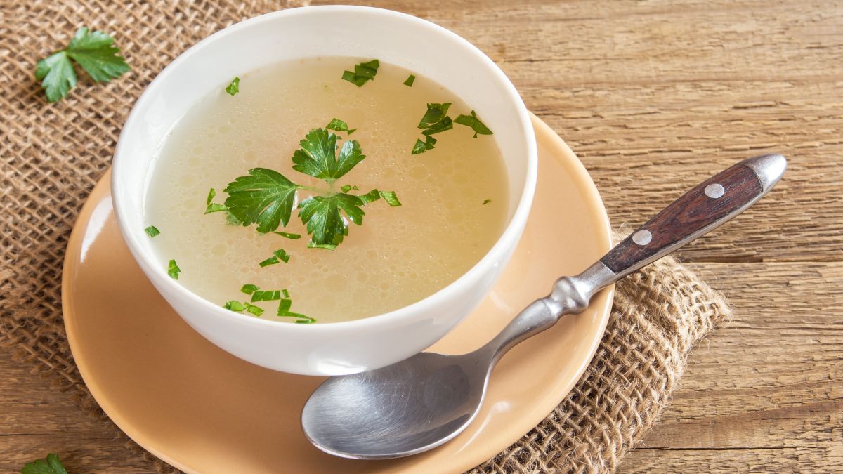 This is a picture of bone broth with parsley in a white bowl on a pale yellow saucer with a silver spoon on a wooden table background