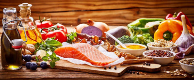 This is a picture of many Mediterranean diet foods such as a salmon filet, olive oils, blueberries, tomatoes on the vine, bell peppers, broccoli, onions, and turmeric powder on a wooden table in glas bottles, 