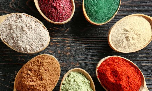 This is a picture of 7 wooden spoons each containign a different kind and color of superfood, on a dark wooden background.