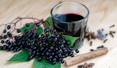 This is a picture of freshly picked elderberries next to a glass of elderberry juice in a glass cup on a wooden tabletop.