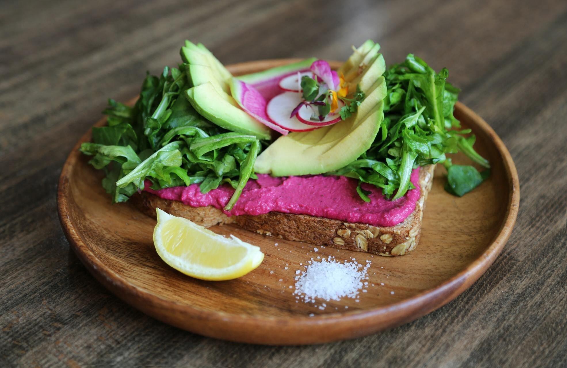 This is a picture of wheat toast on a wooden plate topped with Beet Root Hummus, avocado slices, radishes and greens, with lemons and salt alongside, all on a wooden background.