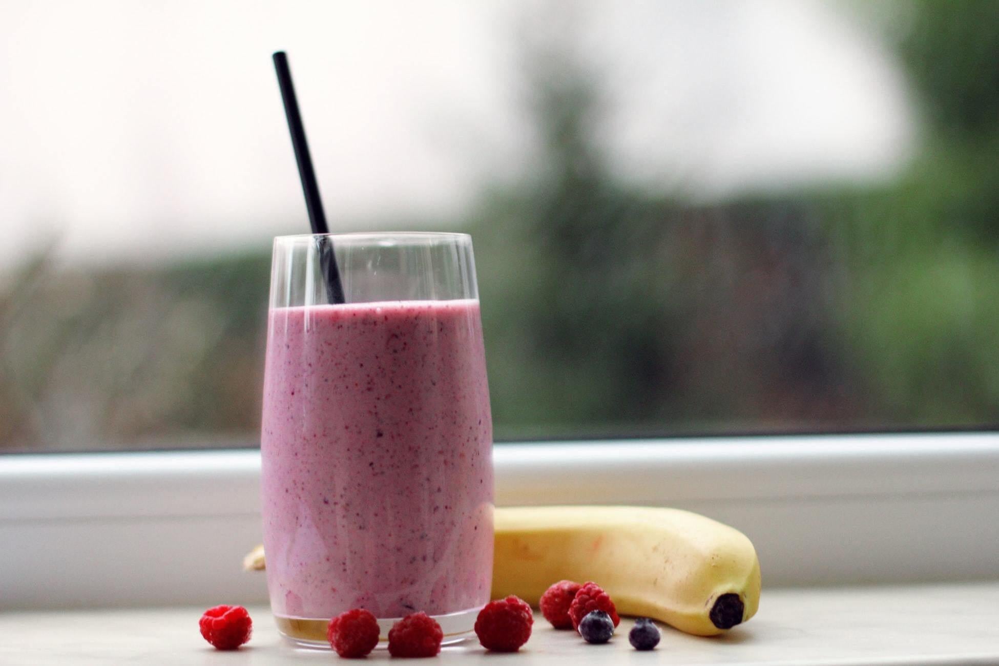 This is a picture of our Berry Collagen Peptide Detox Smoothie in a glass jar wit ha black straw on a white countertop with an outside blurred background.