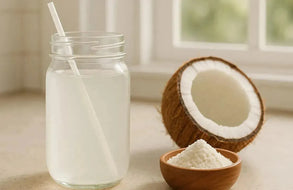 This is a picture of a glass of coconut water in a mason jar with a white straw next to a half a coconut and a coconut water powder in a wooden bowl, on a kitchen counter next to a window.