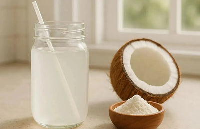 This is a picture of a glass of coconut water in a mason jar with a white straw next to a half a coconut and a coconut water powder in a wooden bowl, on a kitchen counter next to a window.