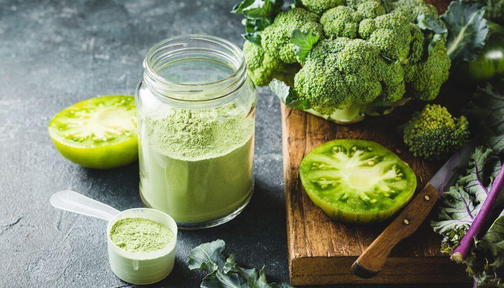 This is a picture of a green smoothie in a glass jar next to green vegetables on a wooden cutting board.