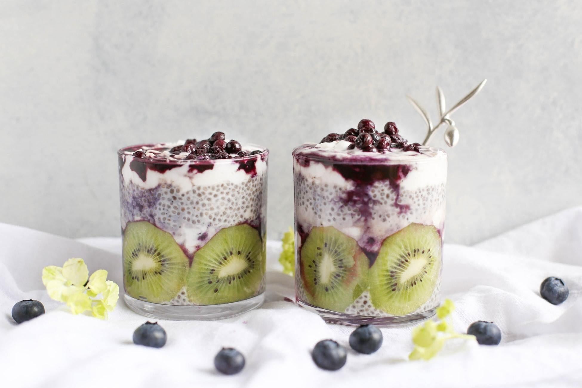 This is a picture of two glass cups filled with Chocolate and Banana Chia Seeds Pudding with Almonds with blueberries sprinkled around them on a white background.