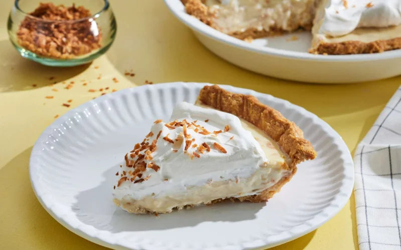 This is picture of a slice of our Coconut Cream Pie with a fork in the middle on a white plate with a white background.