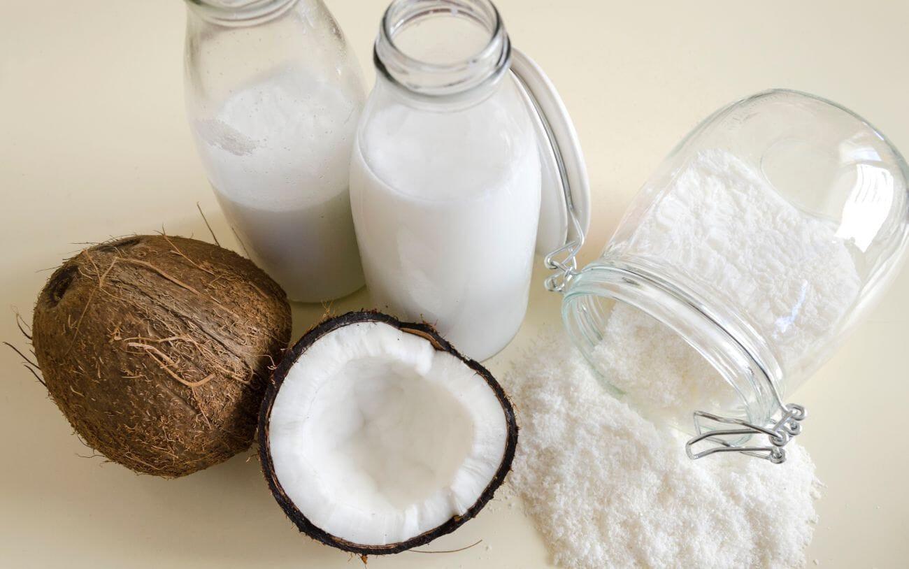 This is a picture of two glass jars with coconut milk and a glass jar on its side with coconut milk powder, next to two coconuts with a light background.