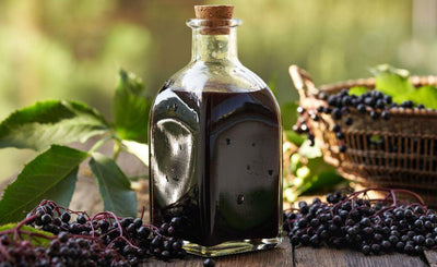 This is a picture of elderberry juice in a glass container with a fresh elderberries resting next to it on a wooden table top.