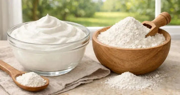 This is a picture of a glass bowl full of fresh sour cream next to a bowl of sour cream powder on a kitchen counter, with a window overlooking a sunny day with fresh trees and grass in the backyard.