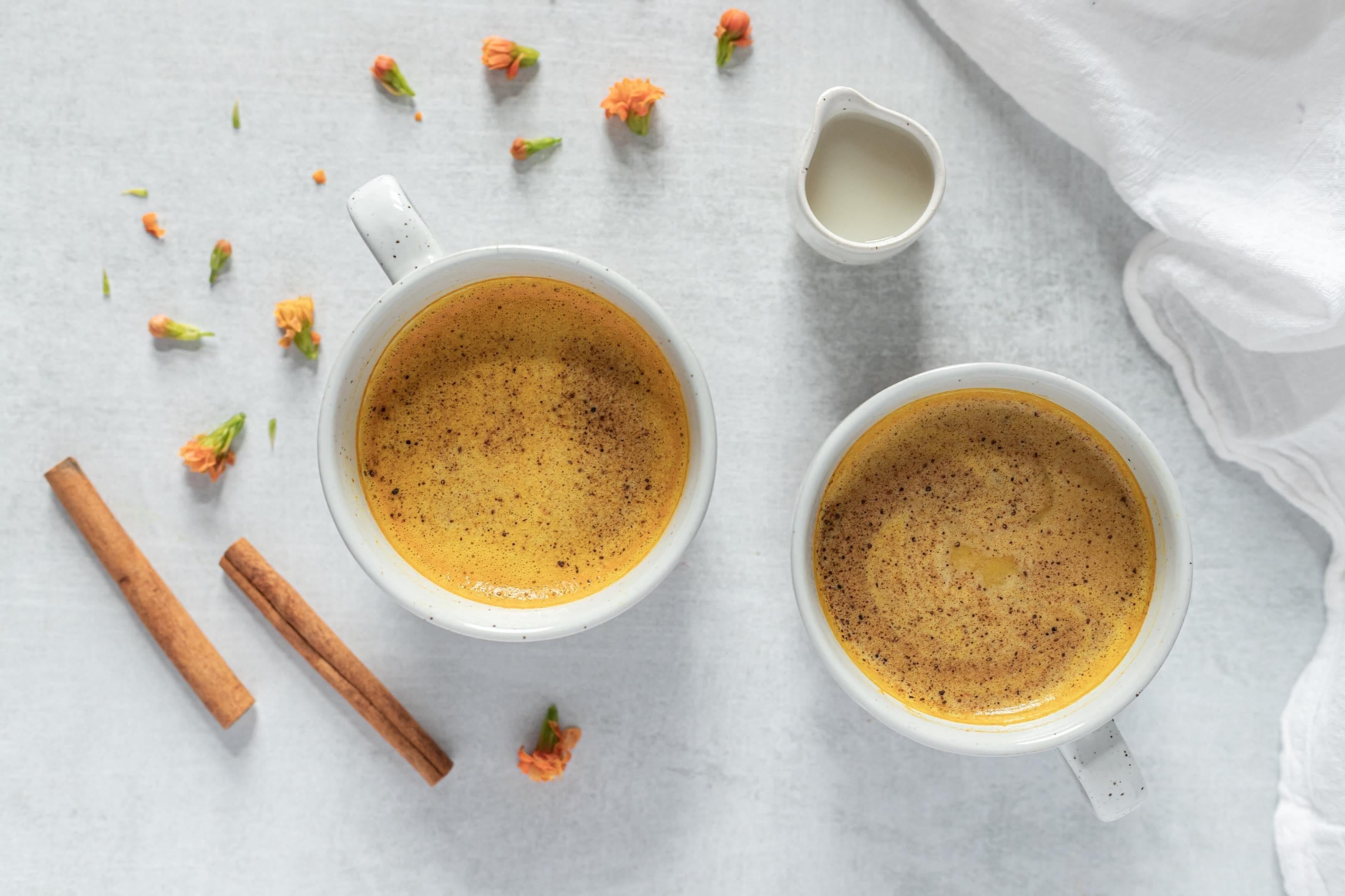 This is a picture of two white mugs full of Golden Milk with cinnamon sticks and herbs off to the side on a white tablecloth.