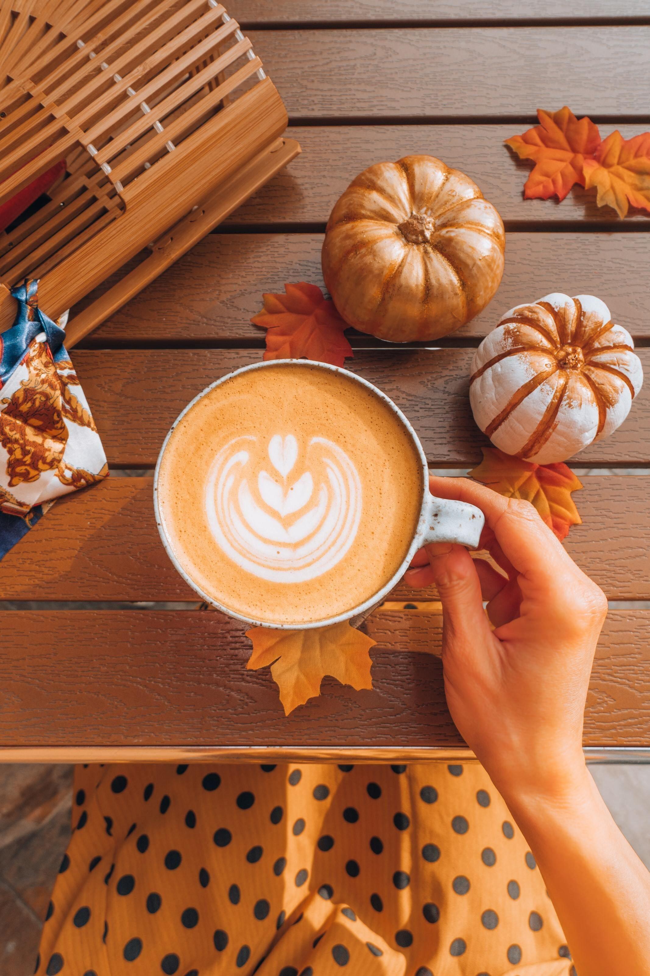 This is a link of our Pumpkin Spiced Latte in a ceramic mug on a wooden table with pumpkins and leaves on the table.