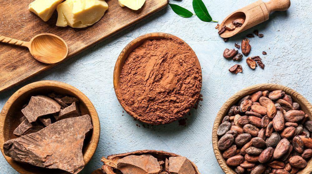 This is a picture of cacao powder, cacao nibs and chocolate in wooden bowls on a light background.