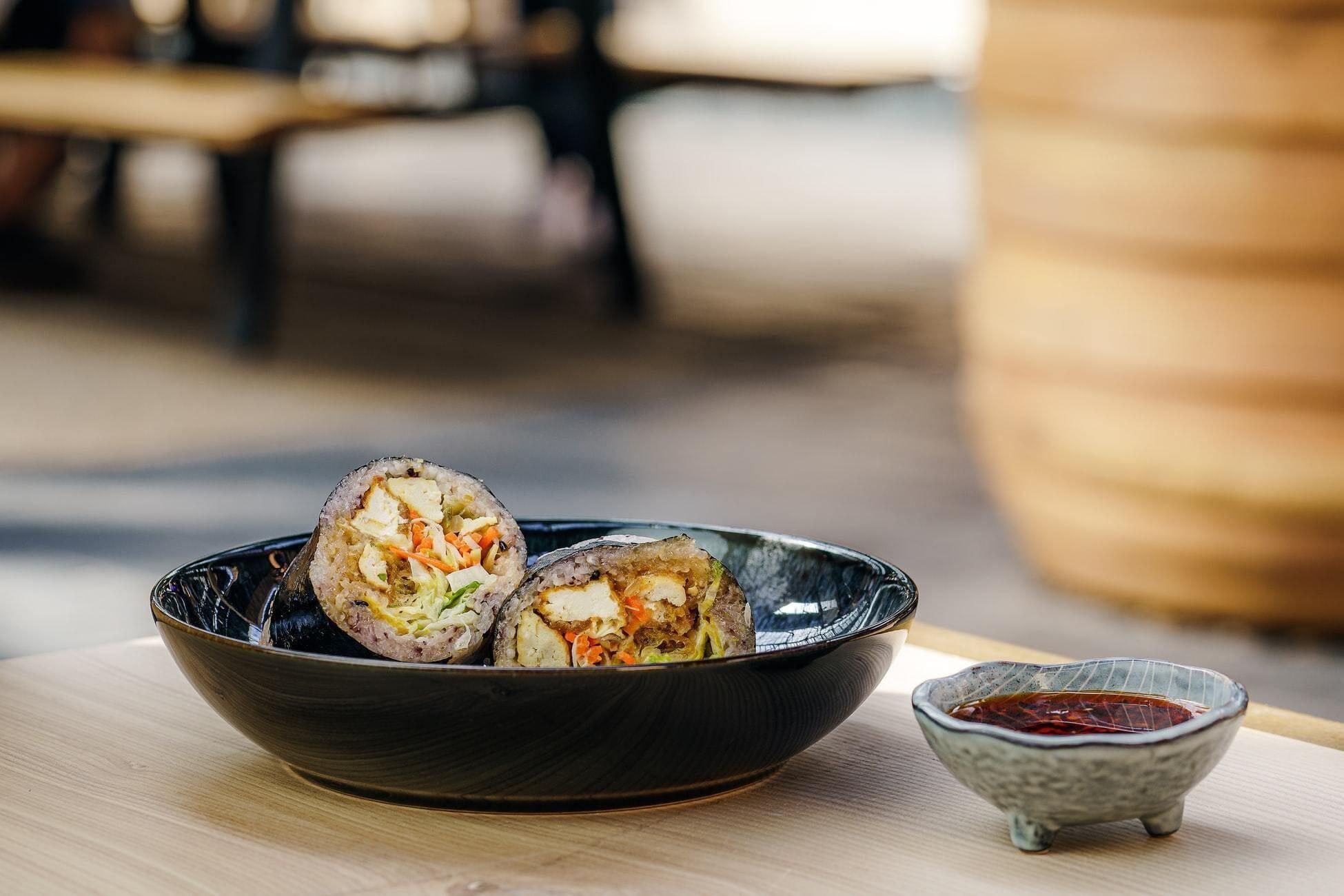 This is a picture of our Kale Marinated Tofu Wrap in a black bowl with a side of sauce on a countertop with a restaurant style setting in a blurred background.