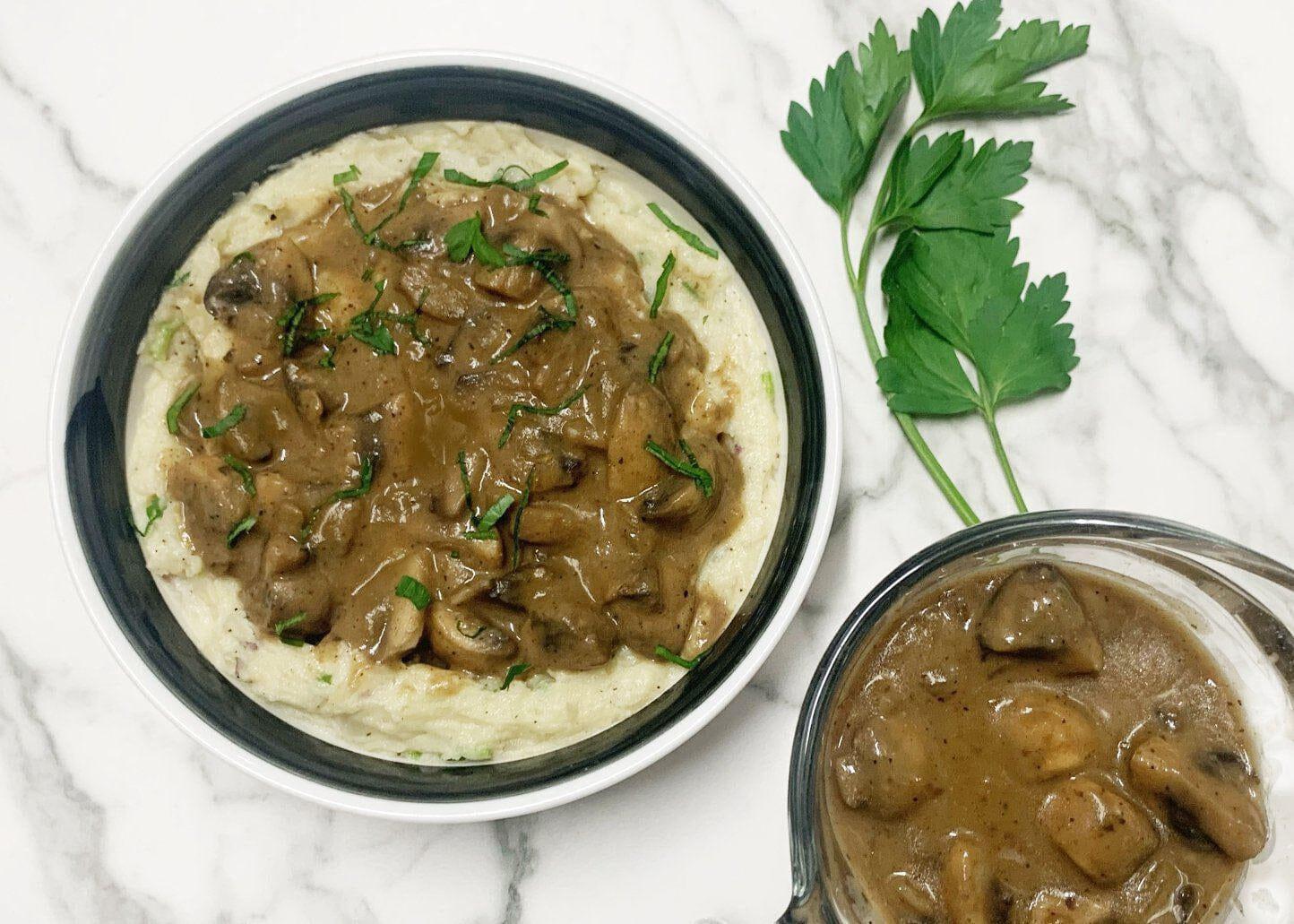 This is a picture of Mashed Potatoes with Vegan Mushroom Gravy in a ceramic bowl, and a side of gravy in another ceramic bowl on a marble table.