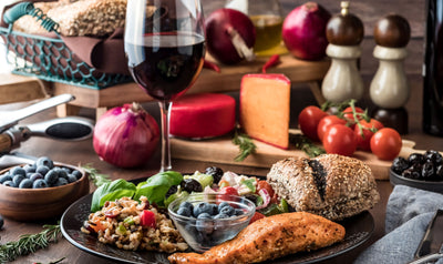 This is a picture of many foods in the Mediterranean diet including a salmon filet on a plat with rice and blueberries and vegetables, with a bread roll and a glass of red wine., with cheese, tomatoes, an onion and spices in the background.