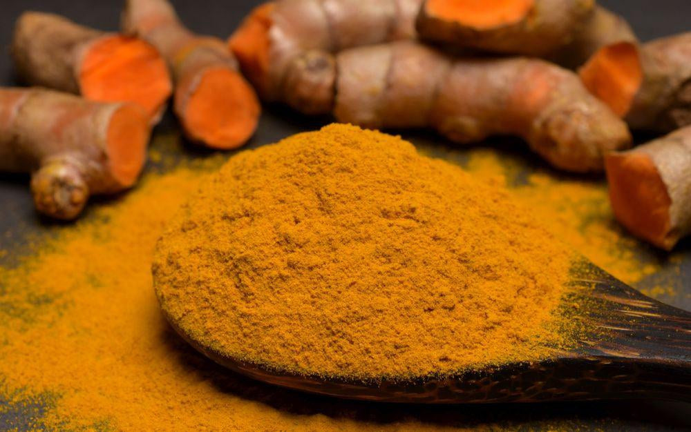 This is a picture of fresh cut turmeric root pieces around a wooden spoon filled with turmeric powder on a wooden table top