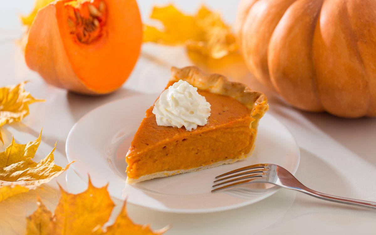 This is a picture of a slice of Pumpkin Pie with whip cream on top on a white plate, with 3 pine cones in the background on a wooden table top.