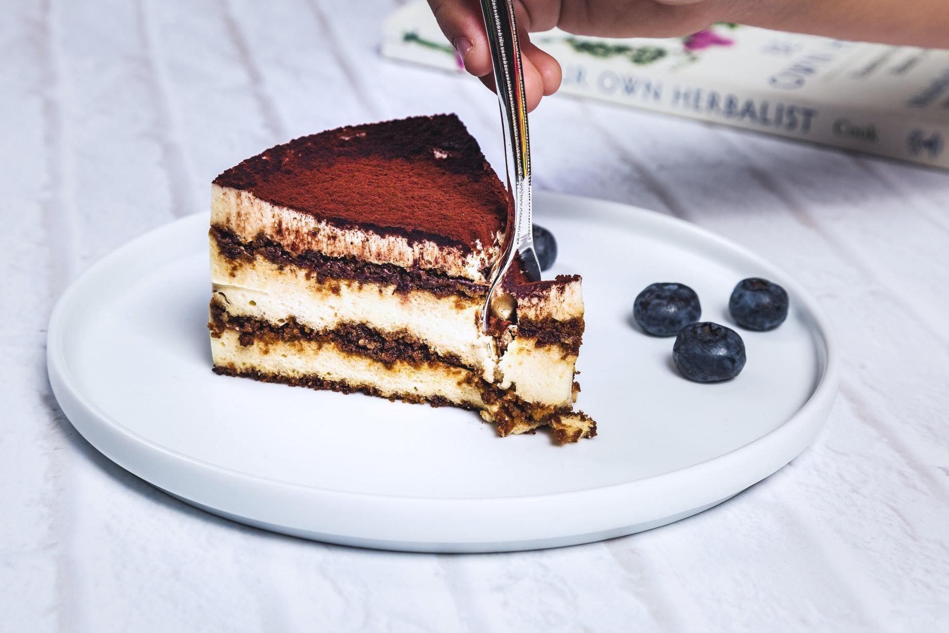 This is a picture of a delicious power protein vanilla-coconut cake on a white plate with a fork slicing into it, with 3 blueberries resting next to it.