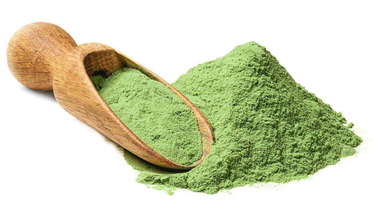 This is a picture of a green powder next to a large wooden spoon on a white background.