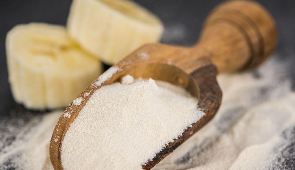 This is a picture of a wooden spoon filled with banana powder  with sliced bananas behind it