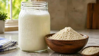 This is a picture of a close up of a mason jar filled with delicious fresh buttermilk next to a wooden bowl filled with buttermilk powder on a counter near a kitchen window.