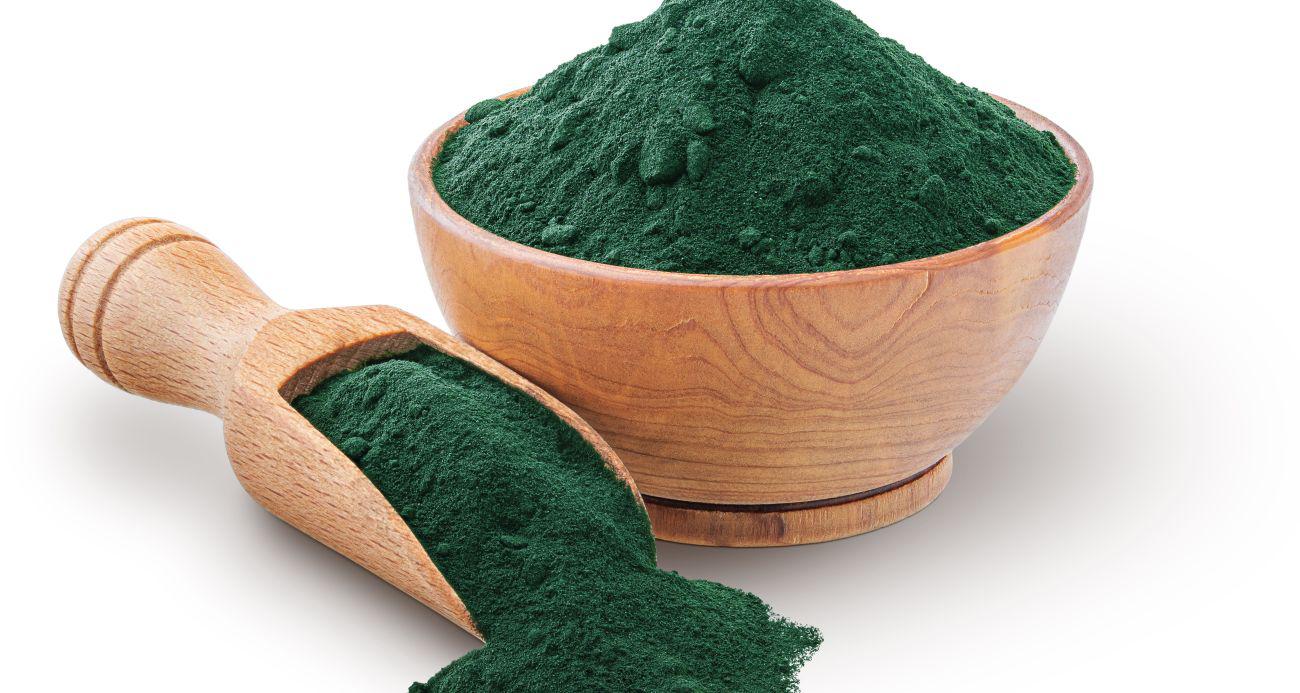 This is a picture of vibrant green spirulina powder in a wooden bowl, with a large wooden spoon next to it will ed with spirulina powder on a while background. 