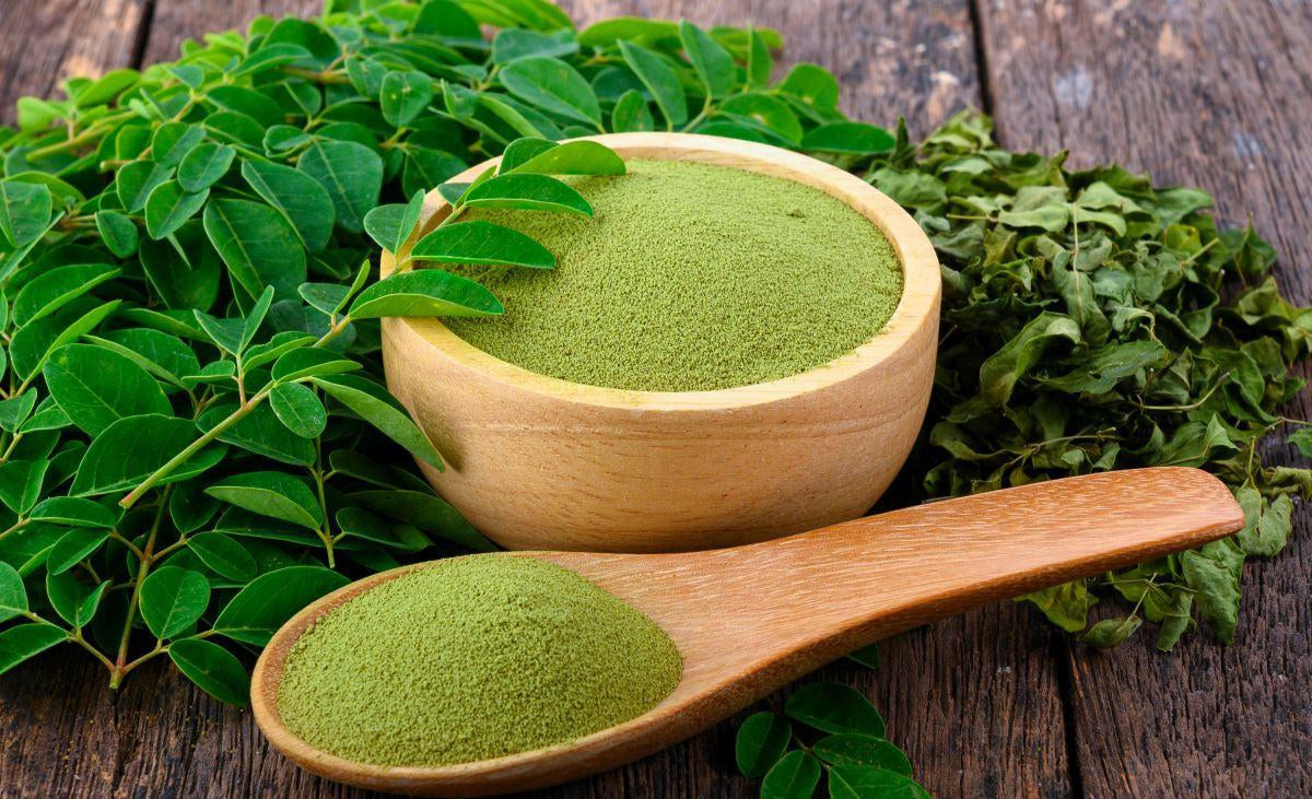 This is a picture of a wooden bowl and a wooden spoon filled with Moringa powder next to moringa leaves on a wooden table.