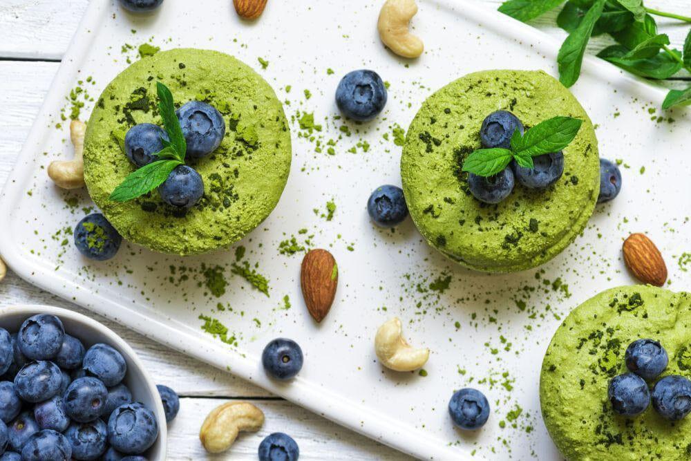 This is a picture of green superfood blends in glass bowls with blueberries and almonds sprinkled on the table top around them.