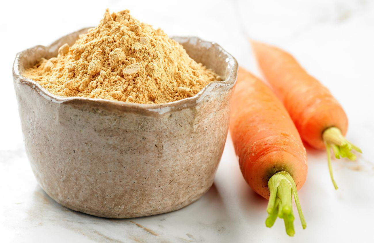 This is a picture of a ceramic bowl of carrot powder with two carrots next to it, on a white tabletop.