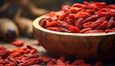 This is a picture of a wooden bowl filled with fresh goji berries on a wooden table, with more goji berries next to the bowl.