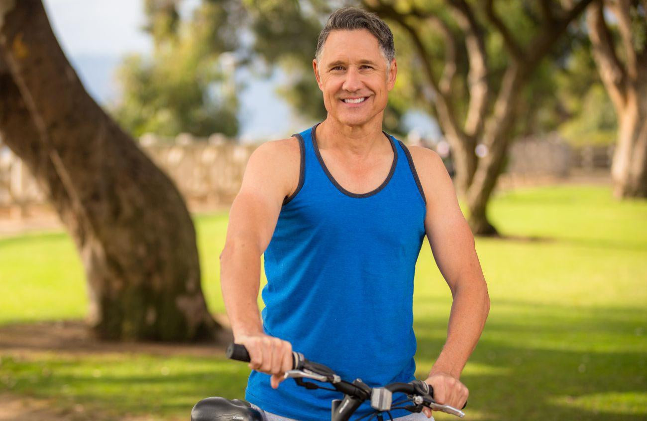 This is an image of a man in a blue tank top with his bicycle in nature with trees in the background