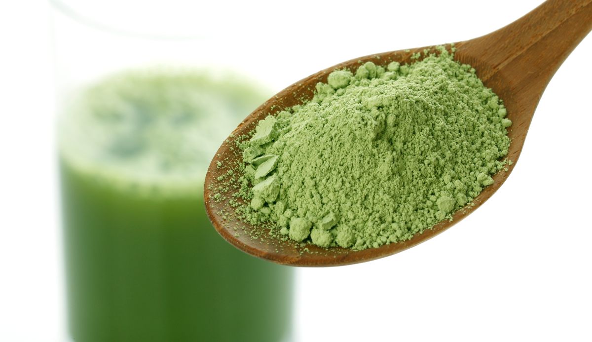 This is a picture of a wooden spoon full of greens powder with a green smoothie in a glass in the background.