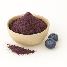 A beige bowl filled with purple blueberry powder on a white background, with a small pile of powder and two fresh blueberries beside it.