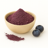 A beige bowl filled with purple blueberry powder on a white background, with a small pile of powder and two fresh blueberries beside it.